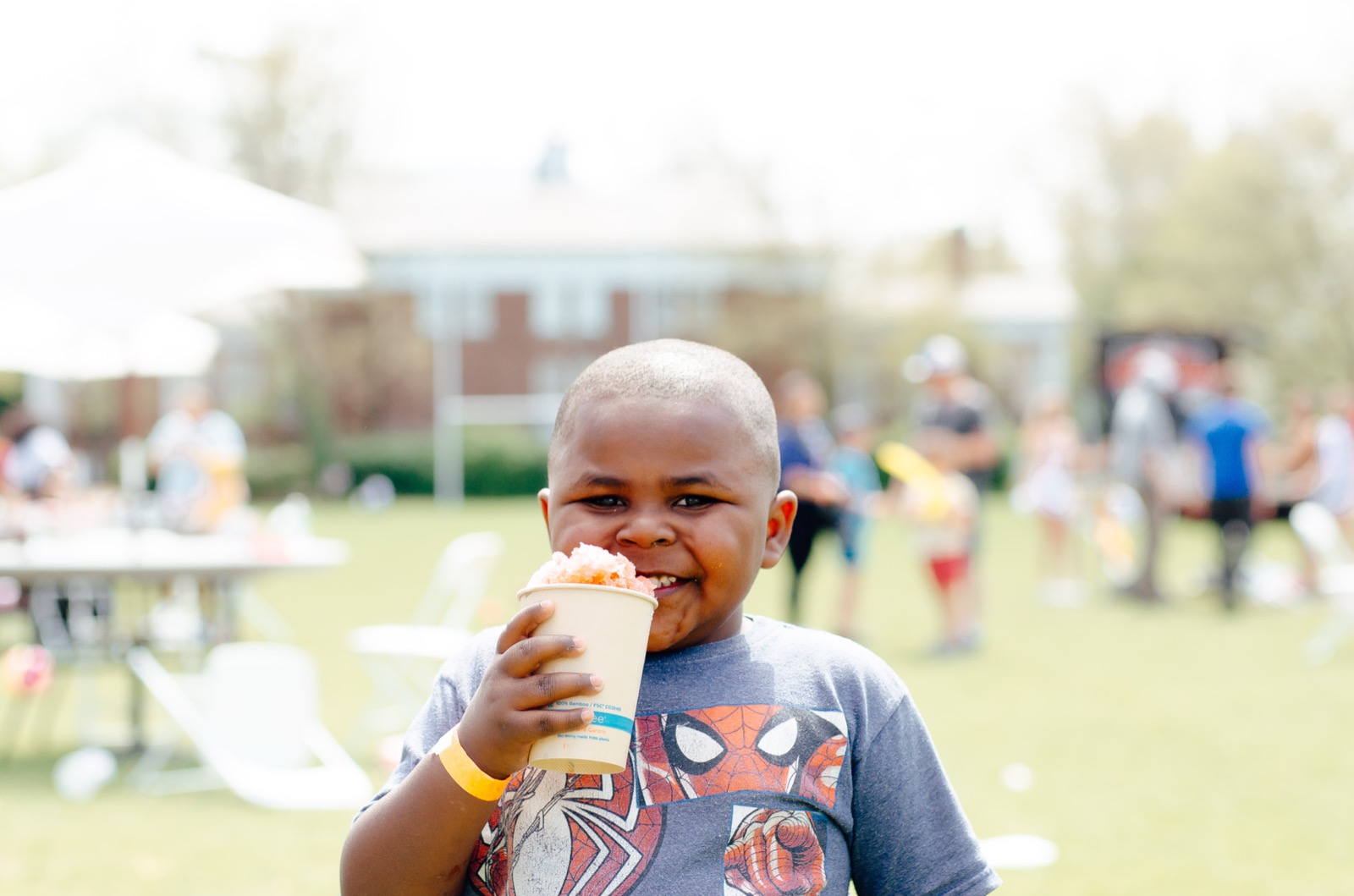 A young child at a City of Promise community event grins behind a paper cup of shaved ice, sunlit lawn behind him.