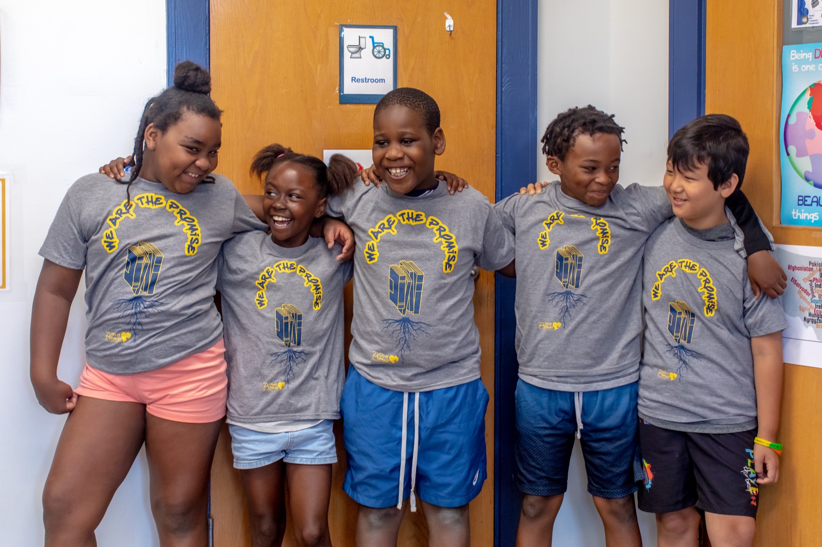 Five City of Promise scholars in matching “We Are The Promise” t-shirts stand arm-in-arm in a school hallway, laughing together.