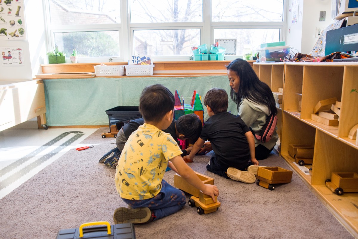 Children at an early learning center play together with wooden toys while a caregiver watches over them.
